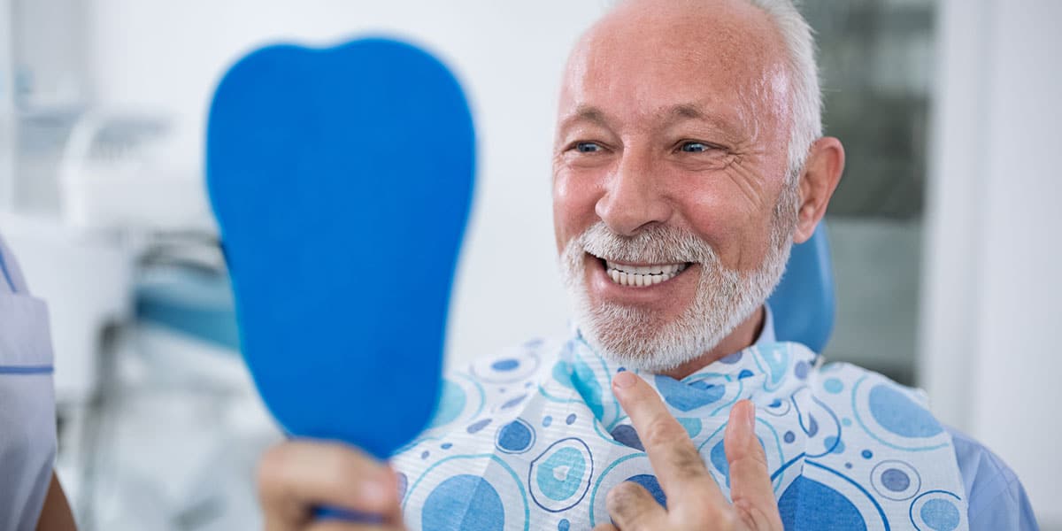 older man in dental chair holding mirror, smiling and pointing at teeth